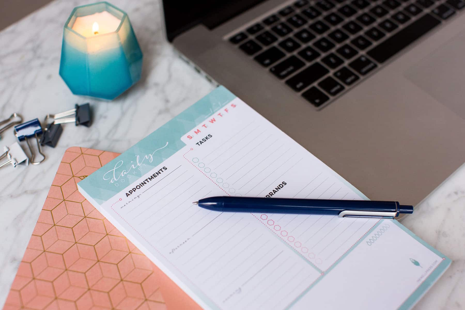 A pen rests atop an Inkwell Press Planner laying next to a macbook pro and a candle. Photographed by Rebecca Marie Art