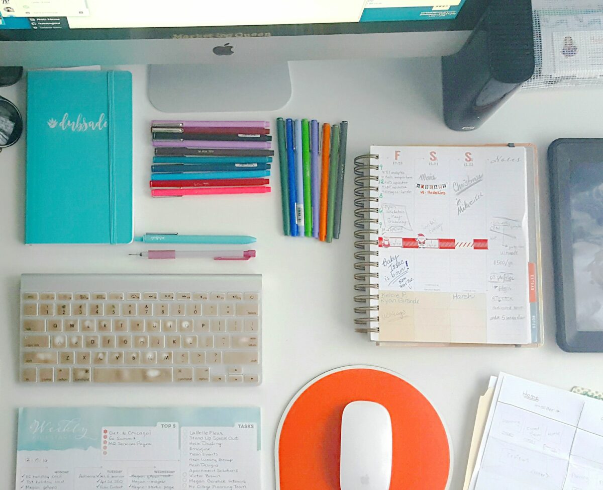A top down view of a desk with many varying office items such as stapler, sunglasses, Inkwell Press Planner, mouse, folders, pens and an iMac computer.