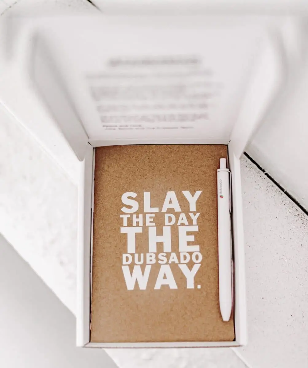 A notebook and pen in an open box. Text on the notebook cover states "Slay the day the Dubsado way"