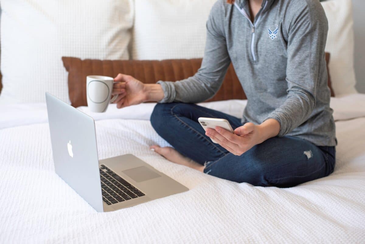 A woman appears to be scrolling on her mobile phone while sitting on a bed with white linens. Her computer is also out and she has a drinking mug in her other hand.