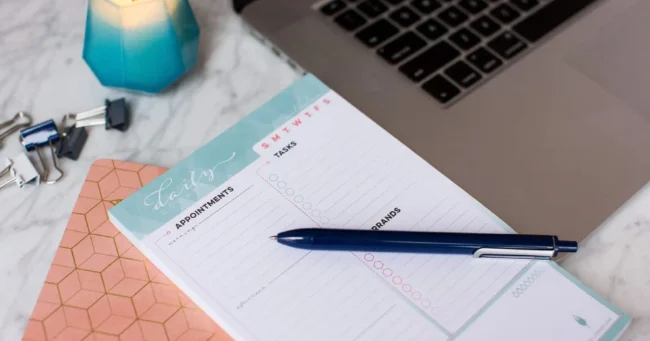 A pen rests atop an Inkwell Press Planner laying next to a macbook pro and a candle. Photographed by Rebecca Marie Art