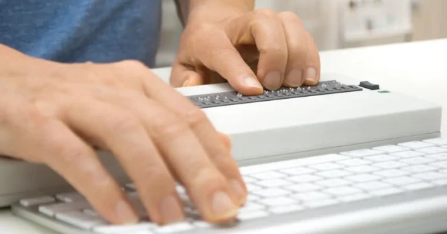 Visually impaired working on computer with assistive technology; braille display and keyboard.
